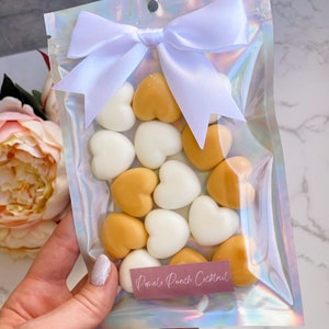 May include: A clear iridescent plastic bag with a white satin ribbon tied in a bow. The bag contains heart-shaped wax melts in white and gold. The label on the bag reads "Pomelo Punch Cocktail".
