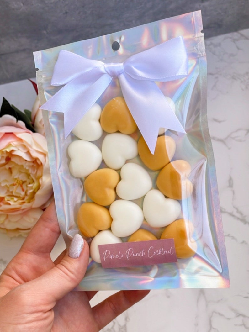 May include: A clear iridescent plastic bag with a white satin ribbon tied in a bow. The bag contains heart-shaped wax melts in white and gold. The label on the bag reads "Pomelo Punch Cocktail".