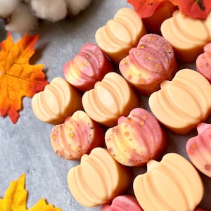 Könnte beinhalten: Draufsicht auf herzförmige Wachsschmelzen in Pfirsich- und Rosatönen, einige mit goldenen Glitzerakzenten. Herbstblätter und Baumwollknospen sind um die Schmelzen angeordnet und erzeugen ein saisonales Thema.