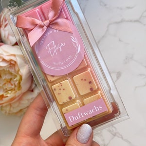 Könnte beinhalten: Ein rosa-weißes Wachs-Schmelzstück in einer durchsichtigen Plastikverpackung mit einer rosa Schleife. Das Wachs-Schmelzstück ist mit "Dufwachs" beschriftet und hat einen Aufkleber mit der Aufschrift "Erstellt von Elisa mit Liebe".