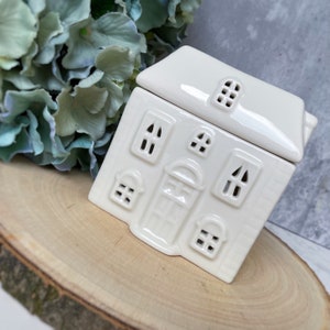 Könnte beinhalten: Ein cremefarbenes Keramik-Haus-Teelichthalter mit abnehmbarem Dach. Das Haus hat geprägte Fenster und eine Eingangstür. Der Teelichthalter steht auf einer Holzoberfläche, mit blau-grünen Blumen im Hintergrund.