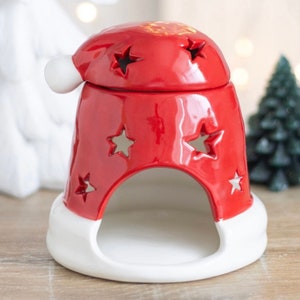Könnte beinhalten: Rote Keramik-Weihnachtsdekoration in Form eines Weihnachtsmützenhuts. Der Hut hat sternförmige Aussparungen und einen weißen Rand. Die Dekoration ist für ein Teelicht oder eine kleine Kerze konzipiert und erzeugt einen festlichen Glanz.