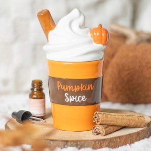 Könnte beinhalten: Ein Keramik-Öldiffusor in Form eines Pumpkin Spice Latte mit weißer Schlagsahne, einem kleinen orangefarbenen Kürbis und einem Zimtstange. Der Becher ist orange mit einem braunen Band mit der Aufschrift "Pumpkin Spice".