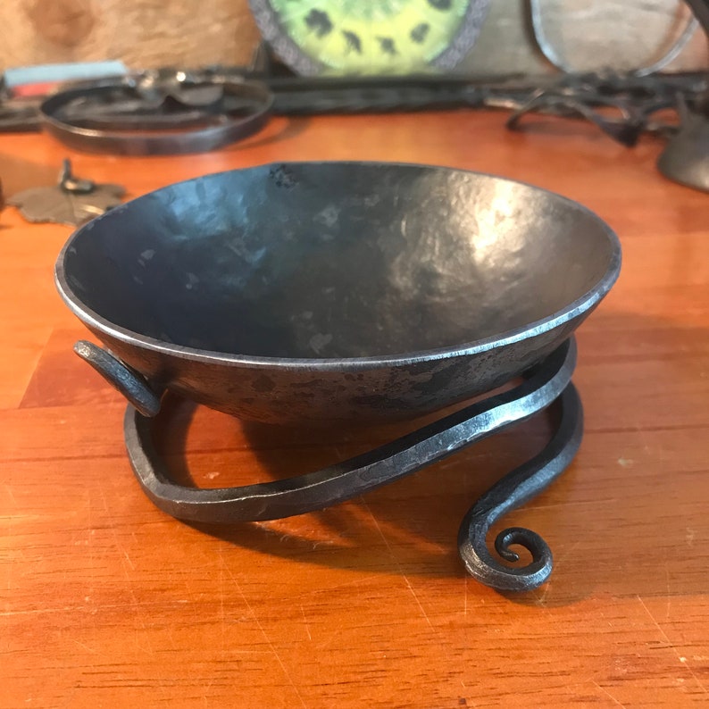 May include: A small, black metal bowl with a hammered finish sits on a curved, black metal stand. The stand has a spiral design at the base.