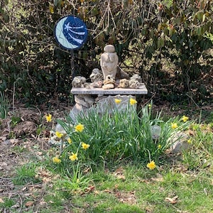 May include: A small stone sculpture of a woman with a round head and a large bust, surrounded by rocks and shells, sits on a stone platform. A blue and white mosaic circle with an arrow design is in the background.