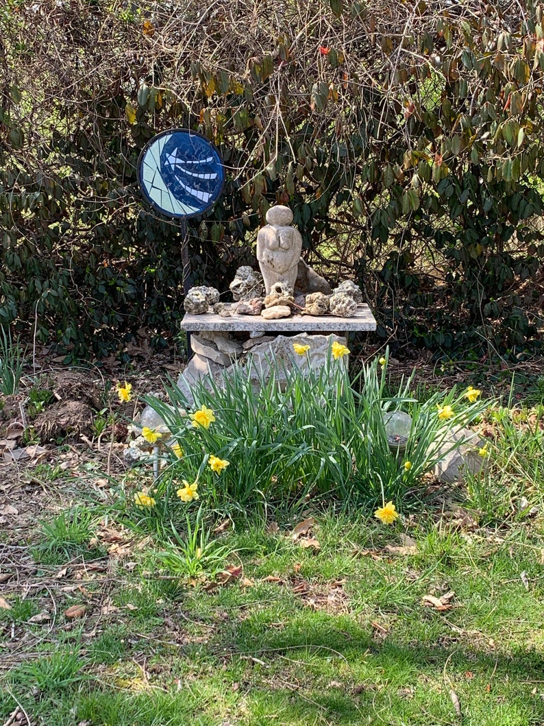 May include: A small stone sculpture of a woman with a round head and a large bust, surrounded by rocks and shells, sits on a stone platform. A blue and white mosaic circle with an arrow design is in the background.