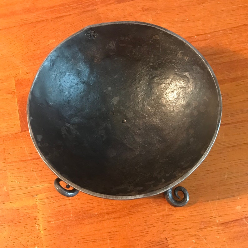 May include: A black metal bowl with three curled legs. The bowl is made of a hammered metal and has a smooth, slightly textured surface.