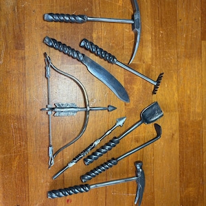 May include: A collection of handcrafted metal tools, including a pickaxe, rake, hammer, and trowel, arranged on a wooden surface. The tools have twisted handles and are likely for gardening or crafting.