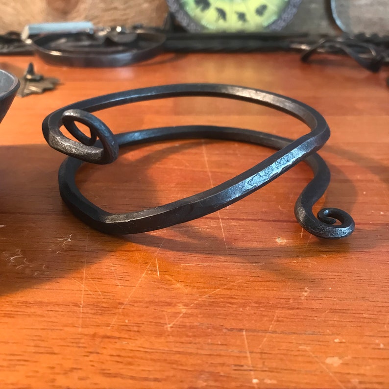May include: A simple, hand-forged metal bracelet with a spiral design at each end. The bracelet is made of dark metal and has a rustic, handmade look.