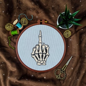 Peut inclure: Une main de squelette blanche faisant un doigt d'honneur est brodée sur un tissu bleu clair tendu sur un cercle de broderie en bois. Le cercle est sur une surface brune avec une petite plante en pot, du fil et des ciseaux.