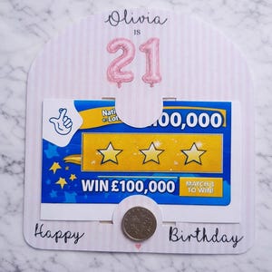 May include: A birthday card with the name "Olivia" and the number "21" in pink. The card includes a lottery ticket design with the text "WIN £100,000" and the words "Happy Birthday". A coin is placed at the bottom.