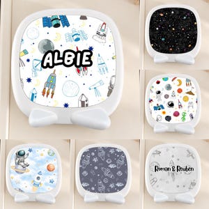 May include: A collection of six square white plastic desktop phone stands, each featuring a unique space-themed graphic print. Designs include rockets, planets, astronauts, and stars, with some featuring the names Albie or Roman & Reuben.
