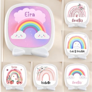 May include: A collection of white, square-shaped vanity mirrors with rounded corners and integrated stands. Each mirror features a unique, colorful graphic print of rainbows, clouds, flowers, or strawberries, along with personalized names like Eira, Amelia, Evie, Freddie, Lucia, and Isabelle.