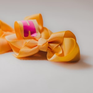 May include: A yellow bow with a pink letter "A" on it. The bow is made of satin ribbon and is tied in a simple knot.