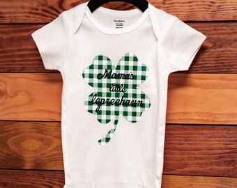 May include: White baby onesie with a green and white checkered shamrock design. The shamrock has the text "Mama's little leprechaun" on it.