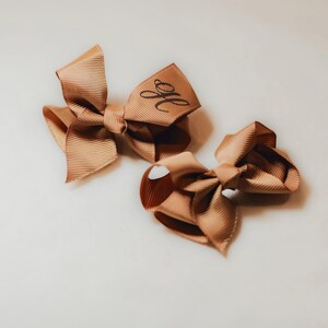 May include: Two brown hair bows with a monogrammed initial "H" on one bow.