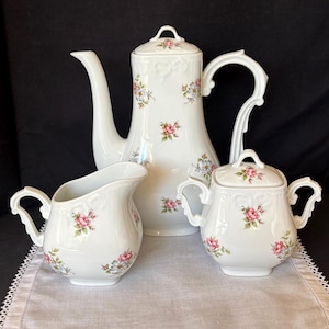 May include: A white porcelain tea set consisting of a tall teapot with a lid, a creamer, and a lidded sugar bowl. Each piece features a delicate floral pattern with small pink roses and light blue flowers scattered across the smooth surface.