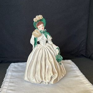 May include: A ceramic figurine of a woman in a white dress with a green jacket and hat. The hat is decorated with gold flowers. The woman is holding a green purse.