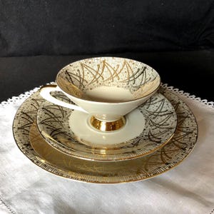 May include: A cream-colored porcelain tea cup, saucer, and small plate set featuring a repeating gold and black abstract geometric pattern. The cup has a gold-toned base and handle, while the saucer and plate have matching gold-toned rims.