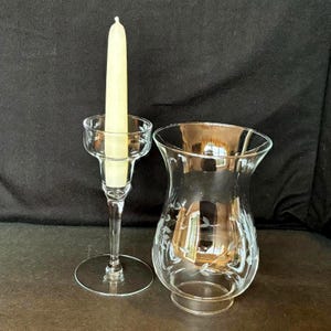 May include: A clear glass candlestick with a lit, cream-colored candle and a clear glass hurricane lamp with etched floral designs. The candle holder has a slender stem and a round base. The hurricane lamp has a flared top and a round base.