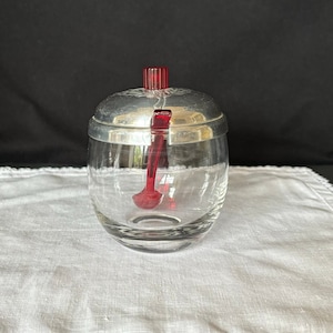 May include: A clear glass sugar dispenser with a silver lid and a red plastic spoon. The lid has a red knob on top.