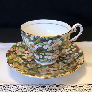 May include: A white teacup and saucer with a floral pattern. The pattern features white flowers with yellow centers on a green background with red accents. The cup and saucer have a gold trim.