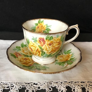 May include: A white teacup and saucer set featuring yellow roses with green leaves. The teacup has a gold handle and trim, and the saucer has a scalloped edge. The floral design is repeated on both pieces, creating a cohesive look.