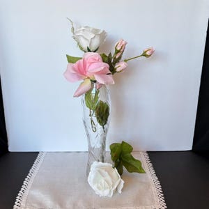 May include: A clear glass vase with a diamond pattern holds pink and white roses and buds. A white rose and green leaves rest on a white lace-trimmed cloth. The vase is on a black surface against a white background.