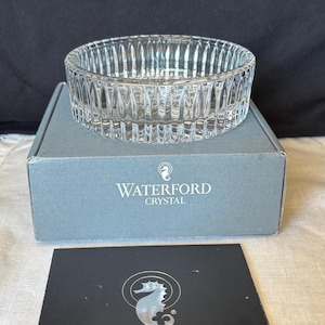 May include: A clear, round crystal bowl featuring vertical cut patterns along its exterior. The bowl rests on a grey cardboard box labeled with the Waterford Crystal logo and a seahorse emblem, accompanied by a black card with a silver seahorse logo.