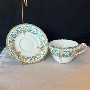 May include: A white teacup and saucer set with a teal, gold, and floral design. The teacup has a scalloped rim and a gold handle. The saucer is displayed on a gold stand. The set is on a light-colored surface.