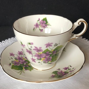 May include: A white teacup and saucer with a floral pattern of purple violets and green leaves. The cup and saucer have a gold trim.