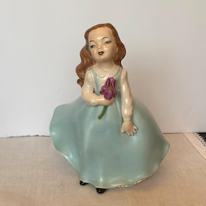 May include: A vintage ceramic figurine of a girl in a light blue dress, holding a purple flower. The figurine has brown hair and is wearing a white blouse. The dress has a full skirt and the figurine is standing.