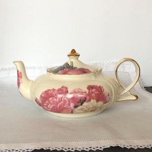 May include: Cream-colored ceramic teapot with a floral design featuring pink roses. The teapot has a gold-colored handle and a gold finial on the lid. The spout and handle are also accented with gold.