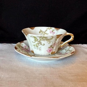 May include: A vintage teacup and saucer set. The white porcelain cup and saucer feature pink roses, green foliage, and gold trim. The cup has a curved handle and a scalloped rim. The saucer mirrors the cup's design.