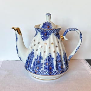 May include: A white porcelain teapot featuring a fluted body decorated with blue floral patterns, a blue checkered base, and gold accents. The teapot includes a matching lid with a knob, a curved handle, and a spout with gold trim.