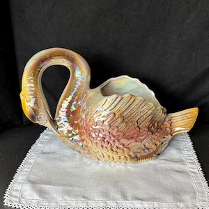 May include: A ceramic swan-shaped planter featuring an iridescent, pearlescent finish with warm golden and bronze tones. The piece displays detailed molded feather textures across its body, a curved neck, and a wide, open top for planting.