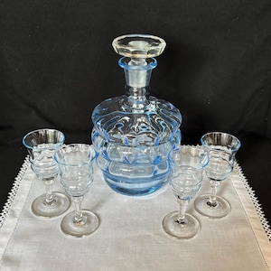 May include: A vintage blue glass decanter set with a stopper and four matching cordial glasses. The decanter has a round, ribbed design. The glasses have a similar ribbed pattern and a stemmed base. The set is displayed on a white cloth.