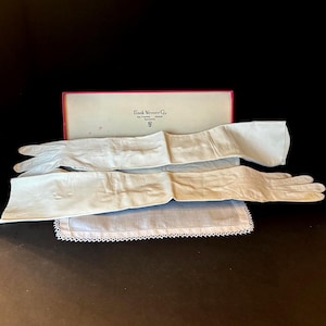 May include: A pair of long, white leather gloves with a matching handkerchief and a box from Frank Werner Co. The gloves are laid out on a dark surface, with the box in the background.