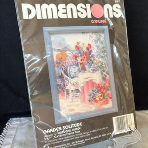 May include: A Dimensions crewel embroidery kit titled "Garden Solitude." The framed image depicts a garden scene with a table, chair, and flowers. The frame size is 5" x 7" (13x18cm). The kit includes cotton embroidery thread and instructions.