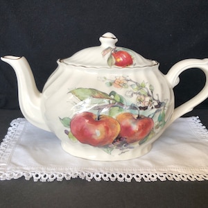 May include: A white ceramic teapot with a floral design featuring apples and blossoms. The teapot has a gold trim around the lid and spout.