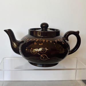 May include: A dark brown ceramic teapot with a lid and handle. The teapot has a rounded body and a curved spout. It is decorated with gold-colored leaf and vine designs.