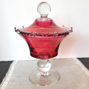 May include: A cranberry red glass candy dish with a matching lid featuring a clear glass ball finial. The bowl has a wide, flared rim with a decorative beaded edge and sits on a clear glass pedestal base with a spherical accent in the center.