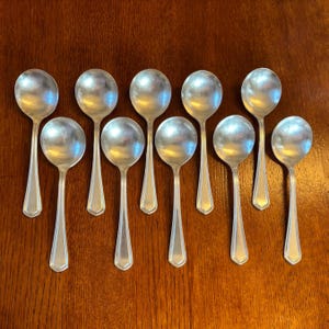 May include: A collection of ten silver soup spoons arranged on a wooden surface. Each spoon features a rounded bowl and a long, slender handle with a decorative, geometric end. The spoons are reflective, with a polished finish.