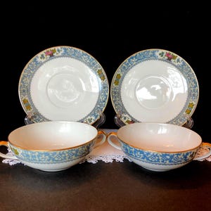May include: Four-piece tea set featuring two cups and saucers. The cups and saucers have a white base with a blue floral border and gold trim. The saucers are round, and the cups have two handles.