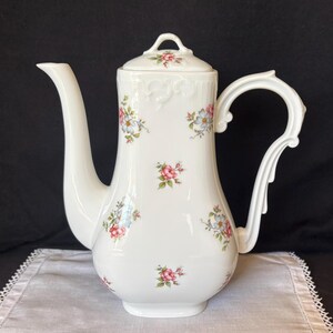 May include: A white porcelain coffee pot featuring a delicate floral pattern with small pink and blue flowers and green leaves. The pot has a tall, slender body, a curved handle, a long spout, and a matching lid with a small floral motif.
