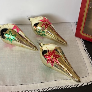 May include: Three vintage Christmas ornaments. The ornaments are teardrop-shaped with a gold finish and cream-colored tops. Each ornament features a red and green floral design. The ornaments are displayed on a white cloth.
