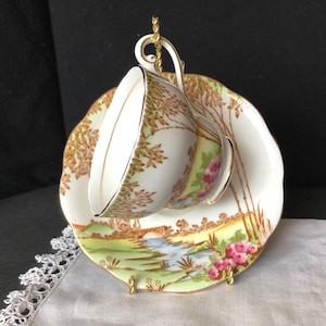 May include: A vintage teacup and saucer set. The white porcelain cup and saucer feature a landscape scene with a bridge, river, and pink flowers. The cup and saucer are trimmed with gold accents. The set is displayed on a gold stand.