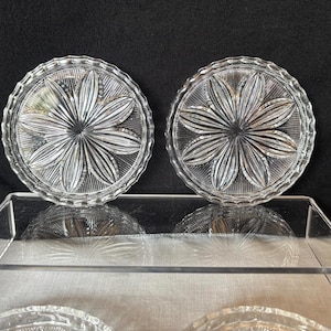 May include: Two clear glass coasters with a flower design. Each coaster has a scalloped edge and a starburst pattern radiating from the center. The coasters are displayed on a clear, reflective surface against a dark background.