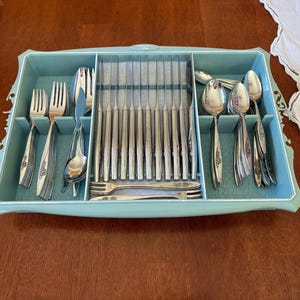 May include: A silver-toned stainless steel flatware set arranged in a light blue plastic storage tray. The set includes forks, spoons, and knives with decorative floral detailing on the handles, organized into three distinct compartments.