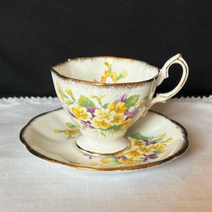 May include: Cream-colored teacup and saucer set with a floral design featuring yellow and purple flowers. The cup and saucer have gold trim. The teacup has a curved handle and sits on a small pedestal base. The saucer is round.